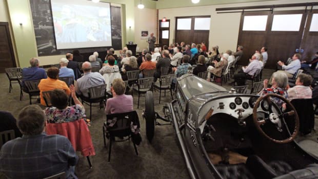  A crowd, sitting amongst a 1916 Packard racer, is enjoying a recent special lecture at the Gilmore Car Museum’s speaker series.