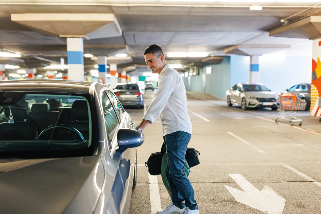 A young Caucasian man is holding a bag in his hand and opening his car door with a smile on his face, while standing in an underground garage.