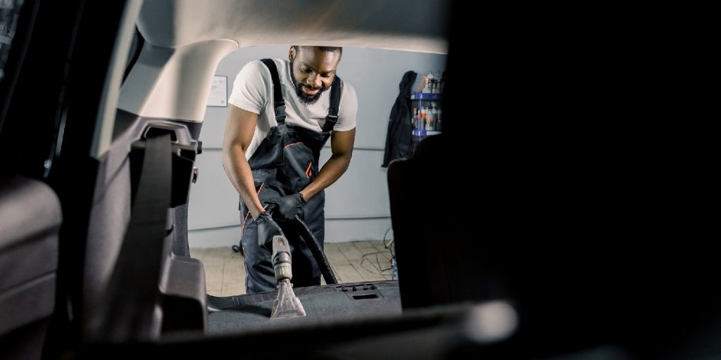 Young African American car wash service worker, doing wet cleaning of luxurious vehicle luggage trunk using vacuum cleaner. View from the car inside.